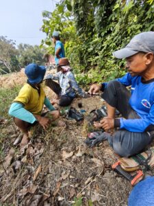 Bang Aep berbaju kuning, Bang Agus berbaju batik, dan Pak Eko berbaju biru duduk bersama di bawah pepohonan di Kebun Balong.